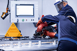 Zwei Leadec-Mitarbeiter in Schutzkleidung bei der Reparatur einer defekten Batterie, auf der linken Seite ein Warnschild für Arbeiten unter Hochspannung.