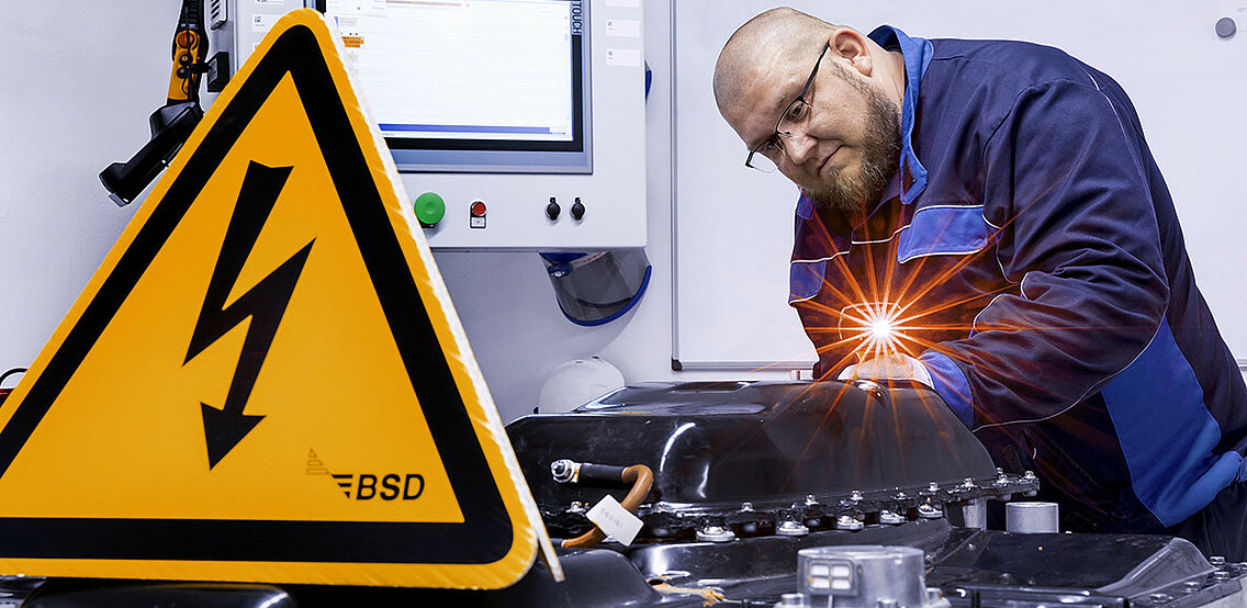 Leadec-Mitarbeiter überprüft eine defekte Batterie, im Vordergrund ein Warnschild für Arbeiten unter Hochspannung.