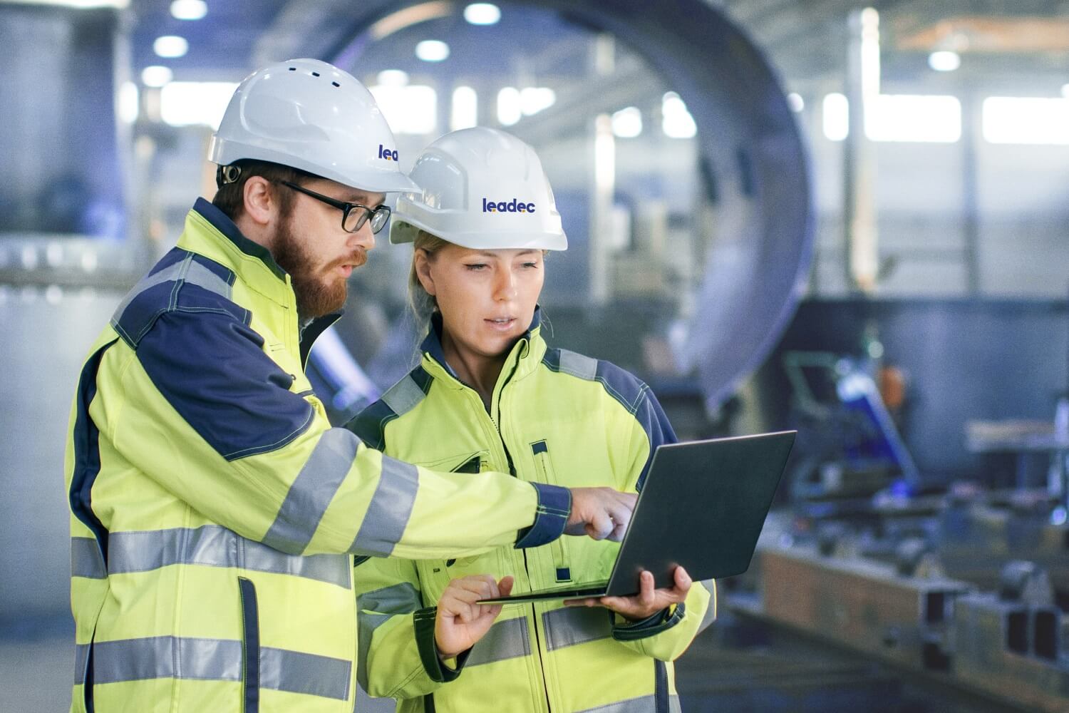 Mann und Frau im Inneren einer Fabrik, die beide Sicherheitskleidung und Helm tragen. 